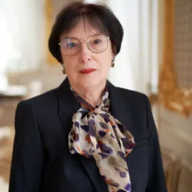 The picture shows a person with dark hair and glasses. She is dressed in a dark jacket with a mottled scarf. She is looking straight ahead, with a slight smile on her face. The background is blurred.