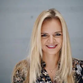The picture shows a person with light hair. She is dressed in a dark T-shirt with bright patterns. She is looking straight ahead, with a smile on her face. The background is subdued.