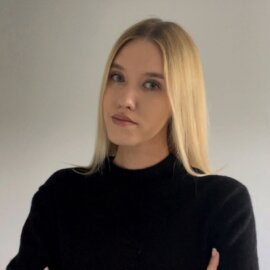 Portret młodej kobiety z blond włosami, ubranej w czarny sweter. Stoi na tle jasnoszarej ściany, z rękami skrzyżowanymi i neutralnym wyrazem twarzy.
