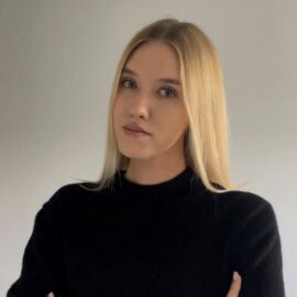 Portrait of a young woman with blonde hair, wearing a black sweater. She stands against a light gray wall with her arms crossed and a neutral facial expression.