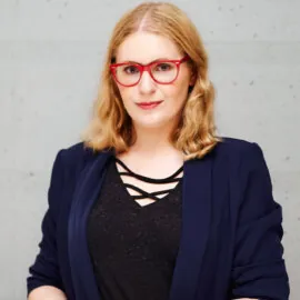 A woman standing against a light gray concrete wall. She has light brown, wavy shoulder-length hair, wears red glasses and delicate makeup. She is dressed in a navy blue jacket, a black blouse with decorative stripes at the neckline and a black watch on her left wrist. She looks straight into the camera with a gentle smile, holding her hands folded in front of her.