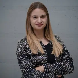 The picture shows a person with light hair. She is dressed in a dark T-shirt and a dark jacket with light patterns. She is looking straight ahead, with a slight smile on her face. The background is subdued.