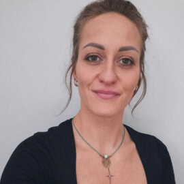 The picture shows a person with brown hair. She is dressed in a dark shirt. She is looking straight ahead, with a slight smile on her face. The background is subdued.