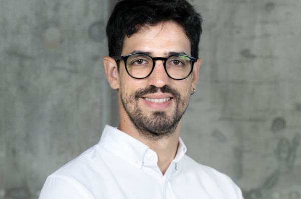 A smiling man with dark hair, a beard, and round glasses stands in front of a concrete wall. He is wearing a white button-up shirt. He appears professional and approachable.