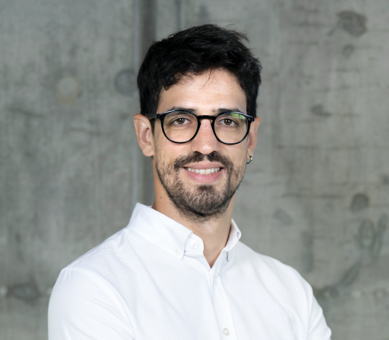 A smiling man with dark hair, a beard, and round glasses stands in front of a concrete wall. He is wearing a white button-up shirt. He appears professional and approachable.
